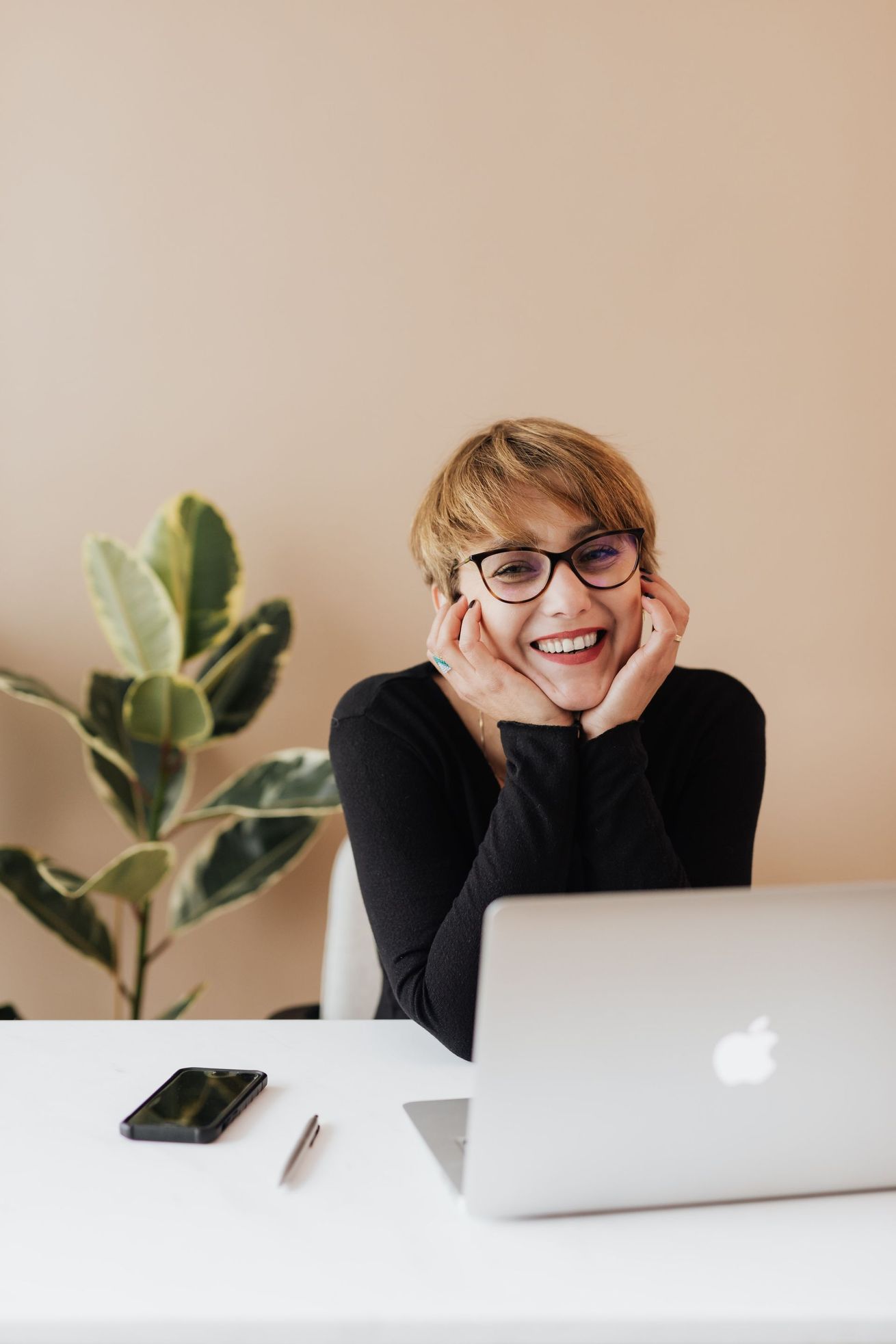 Woman with glasses smiles at the camera, leaning on her hands at a desk with a laptop, phone, and plant.